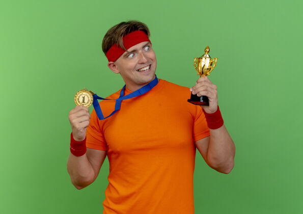 腕带快乐的年轻英俊的运动型男子戴着头带和腕带 脖子上戴着奖牌 拿着奖牌和冠军杯 看着绿色的孤立的杯子持有帅气周围
