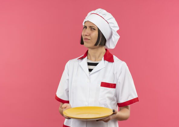 厨师穿着厨师制服的年轻女厨师拿着空盘子 粉红色背景上有复印空间制服盘子年轻
