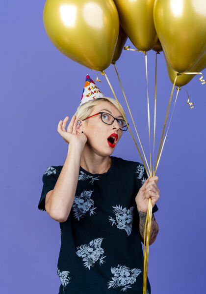 拿着好奇的年轻金发派对女郎戴着眼镜 戴着生日帽 拿着气球在一旁做着听不见你的手势孤立在紫色的墙上生日眼镜人