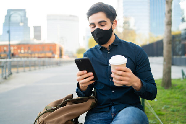 户外年轻人坐在户外的长凳上 一边用手机 一边拿着一杯咖啡保护现代电话