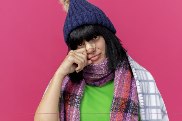 脸特写镜头：戴着冬日帽 围着方格围巾 鼻子上贴着膏药 脸上贴着粉红色墙壁的悲伤的生病的年轻女人公民站围巾