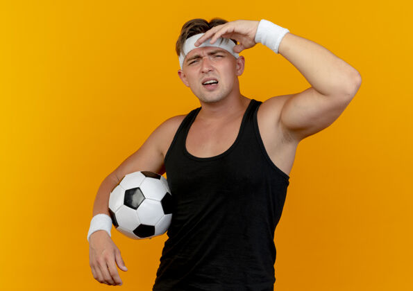 头带年轻帅气的运动型男子戴着头带和腕带拿着足球 把手放在靠近头部的橙色背景上手近抱着