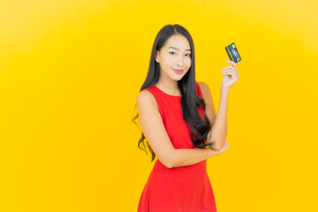 购买画像美丽的亚洲年轻女子微笑着用信用卡在黄色的墙上金融女孩微笑