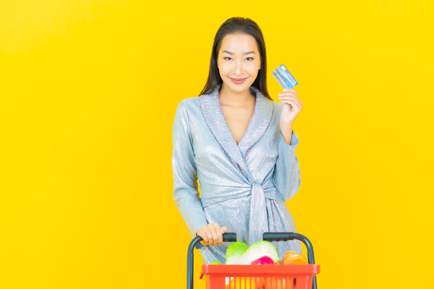肖像一幅美丽的亚洲年轻女子微笑的画像 黄色的墙上挂着超市的购物篮购物车享受信用卡