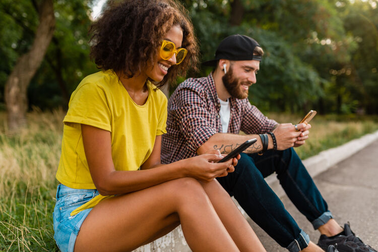 微笑快乐的年轻微笑的朋友坐在公园里用智能手机 男人和女人一起年轻黑色