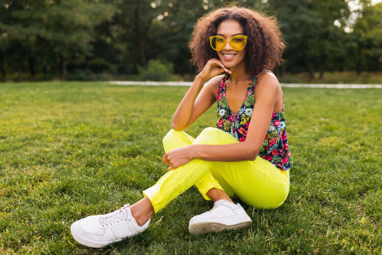 音乐年轻时尚的黑人女子在公园里尽情享受夏日的时尚风格 五颜六色的时髦装扮 坐在草地上戴着黄色的太阳镜和裤子 穿着运动鞋兴奋非洲裔五颜六色