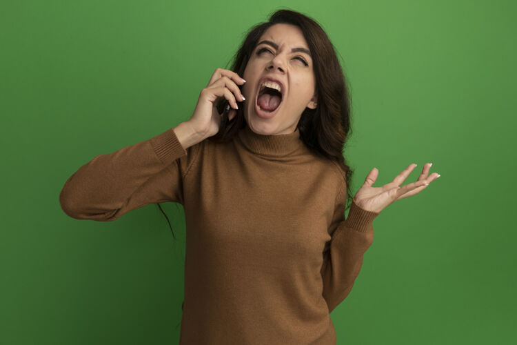 愤怒愤怒的抬头看着年轻漂亮的女孩讲电话 把手孤立地摊开在绿色的墙上电话年轻说话