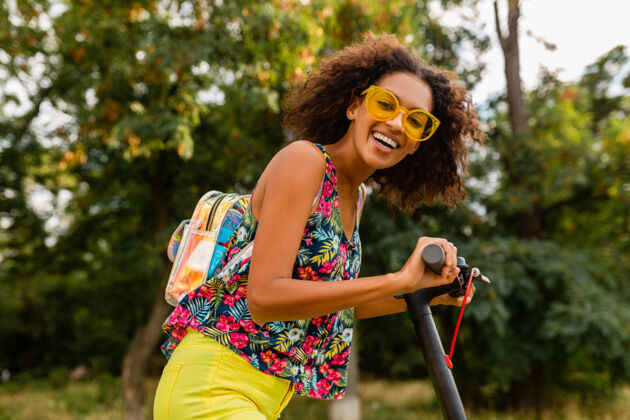 埃斯库特年轻时尚的黑人女子在公园里玩得很开心骑着电动脚踏车在夏天的时尚风格 五颜六色的时髦服装 穿着背包和黄色太阳镜酷微笑环保