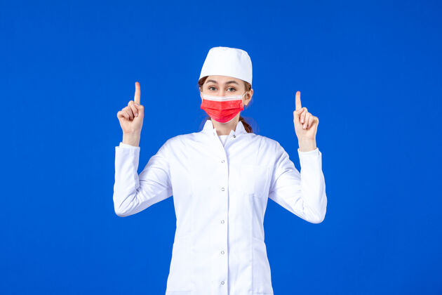 帽子正面图身着医疗服的年轻护士 蓝色墙上戴着红色防护面具医院病毒指向