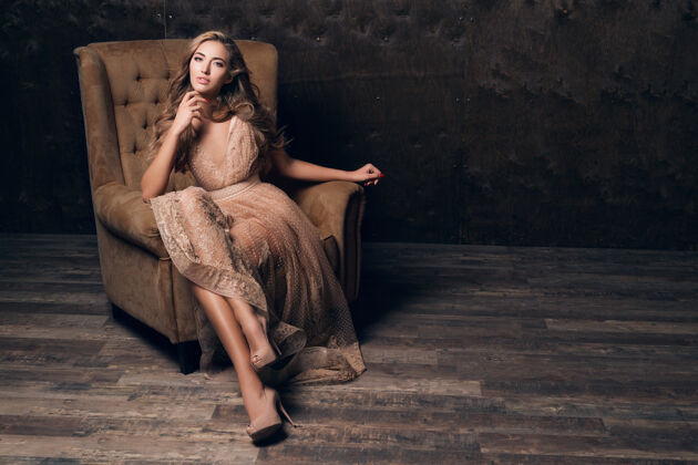夜晚穿着闪亮蕾丝晚礼服的美丽性感模特坐在米色的椅子上华丽年轻女性