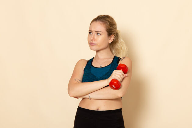 服装正面图身穿运动装的年轻女性手持白色墙壁上的红色哑铃健康运动美体锻炼运动封面女郎身体