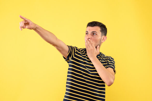 视图正面图穿着黑白条纹t恤的帅哥用手指指着黄色孤立背景上的东西前面手指T恤