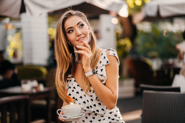休闲美丽的微笑的女人穿着时尚的白色印花连衣裙坐在街上的咖啡馆里喝着一杯卡布奇诺性感姿势咖啡馆