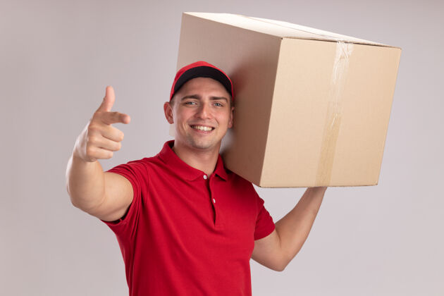 微笑微笑的年轻送货员身穿制服 戴着帽子 肩上扛着一个大箱子 指着隔离在白墙上的摄像机帽子男人制服