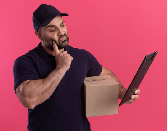 男人印象深刻的中年送货员穿着制服 戴着帽子 拿着盒子 看着手中的剪贴板 把手指放在脸颊上 隔离在粉红色的墙上交货盒子印象