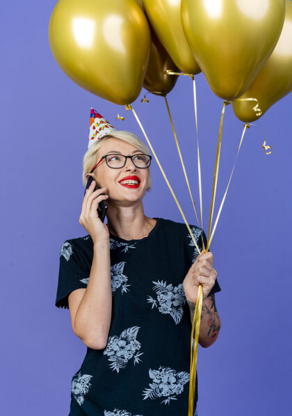 抱着带着微笑的金发派对女孩戴着眼镜 戴着生日帽 手持气球 在紫色背景下孤立地讲着电话紫色生日气球
