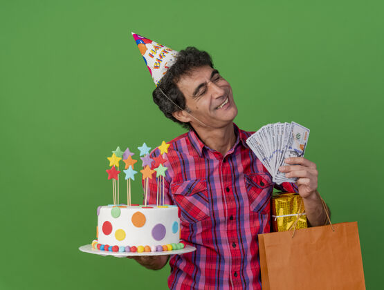眼睛笑容满面的中年白种人聚会男子戴着生日帽拿着生日蛋糕纸袋礼包和钱闭着眼睛隔离在绿色背景和复印空间举行纸中年
