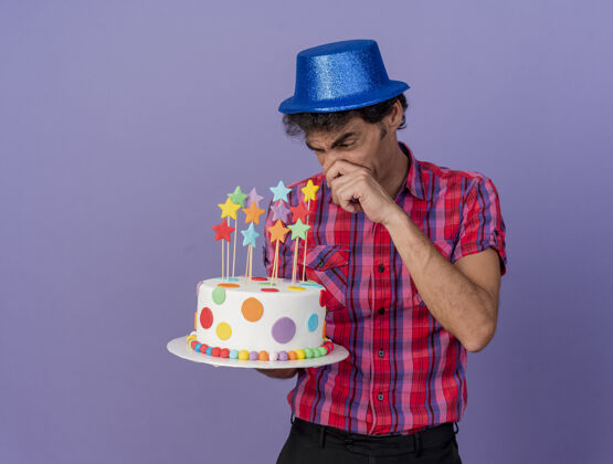 空间恼怒的中年白种人聚会男子戴着聚会帽拿着生日蛋糕看着触摸鼻子的紫色背景与复制空间隔离中年紫色触摸