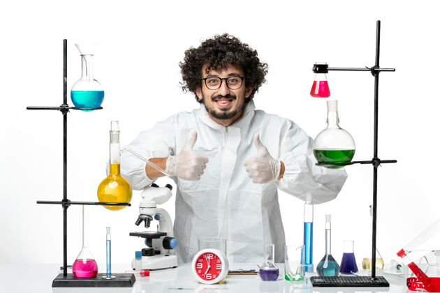 立场正面图身穿特殊套装的年轻男性科学家站在桌子周围 白色的墙上有解决方案烧杯外套特殊