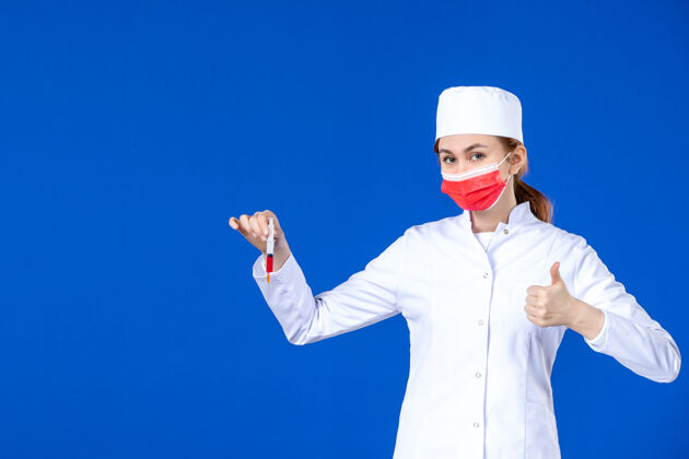 正面正面图：年轻的护士穿着白色的医疗服 戴着红色的面罩 手上拿着针剂 站在蓝色的墙上成人病毒视图