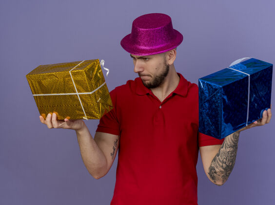 小伙子皱眉年轻英俊的斯拉夫党家伙戴着党的帽子举行 并在紫色背景孤立的礼物包看斯拉夫人打包紫色