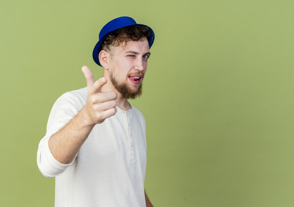 年轻自信的年轻英俊的斯拉夫党家伙戴着党的帽子眨眼显示舌头看和指着相机隔离在橄榄绿的背景与复制空间复制空间帽子