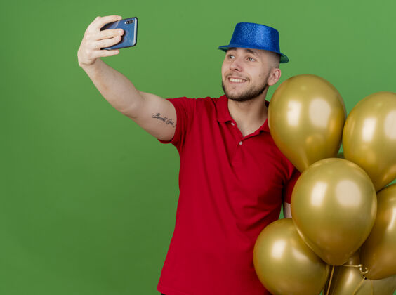 派对微笑的年轻英俊的斯拉夫党人戴着党的帽子拿着气球自拍绿色背景隔离帽子拿着自拍