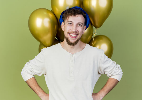 前面面带微笑的年轻英俊的斯拉夫党人戴着党帽站在气球前看着相机手放在腰上橄榄绿的背景隔离斯拉夫派对帽子