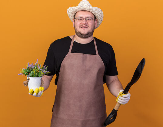 园艺高兴的年轻男园丁戴着园艺帽和手套拿着铁锹 把花盆里的花隔离在橙色的墙上手套帽子请