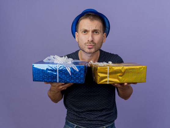 空间无知英俊的白人男子戴着蓝色的帽子拿着礼品盒孤立的紫色背景与复制空间礼物复制紫色
