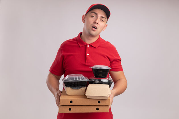 男人疲惫的年轻送货员穿着制服 戴着帽子 把食物容器放在隔离在白墙上的比萨饼盒上帽子盒子送货