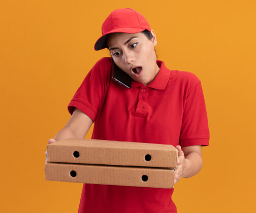 帽子穿着制服 戴着帽子 拿着披萨盒的年轻送货女孩惊讶地说 电话被隔离在橙色的墙上拿着盒子穿