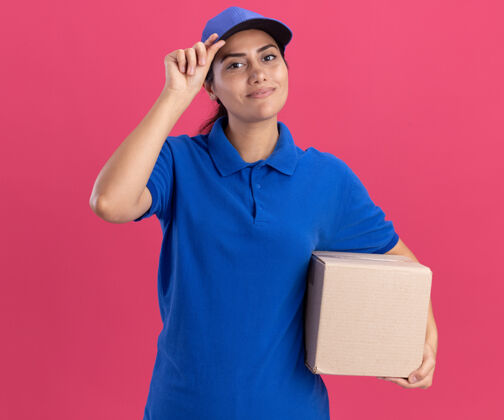 制服年轻的送货员穿着制服 戴着一个装帽子的盒子 帽子被隔离在粉红色的墙上送货戴着盒子