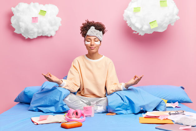 睡眠高兴的非裔美国妇女在床上打坐感到轻松穿着睡衣闭上眼睛练习瑜伽学习后喝咖啡粉色坐着人