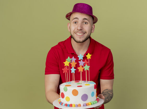 年轻面带微笑的年轻帅哥斯拉夫党人戴着党的帽子拿着生日蛋糕看着相机隔离在橄榄绿的背景与复制空间帽子生日派对