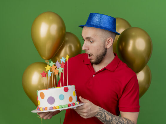 生日惊讶的年轻英俊的斯拉夫党人戴着党的帽子站在气球前拿着生日蛋糕看绿色背景隔离抱着站着斯拉夫