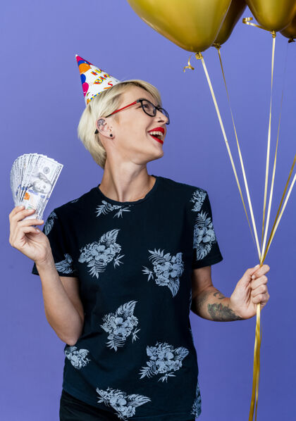 抱着快乐的年轻金发派对女孩戴着眼镜 戴着生日帽 拿着气球和钱 看着紫色背景上孤立的一面钱帽子年轻