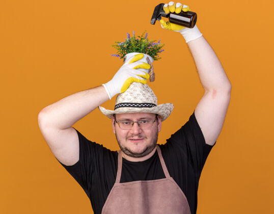 年轻高兴的年轻男园丁戴着园艺帽和手套在花盆里浇花 头上有喷雾瓶隔离在橙色的墙上帽子花穿