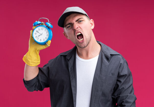 持有愤怒的年轻汉姆森清洁家伙穿着t恤衫 戴着手套拿着闹钟隔离在粉红色的墙上家伙清洁时钟