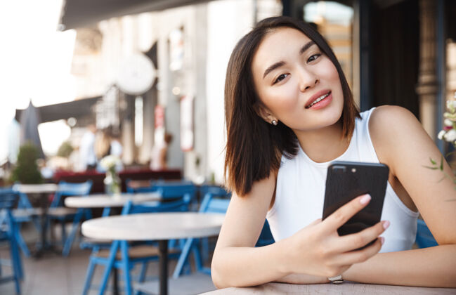 休闲亚洲年轻女子拿着手机坐在咖啡馆里 看着相机神魂颠倒年轻人微笑咖啡