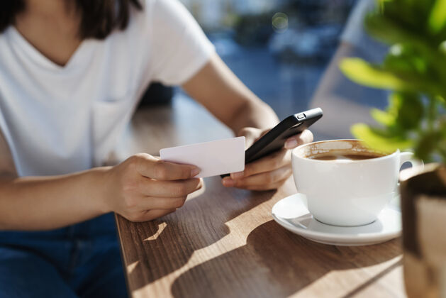 女孩特写镜头：女人的手靠在咖啡桌上 手里拿着手机和塑料信用卡早午餐支付卡布奇诺