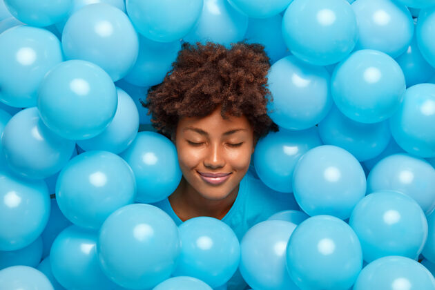 享受高兴的卷发女人闭上眼睛被许多蓝色的充气气球包围有喜庆的心情有乐趣在聚会上感觉很开心成人女性快乐