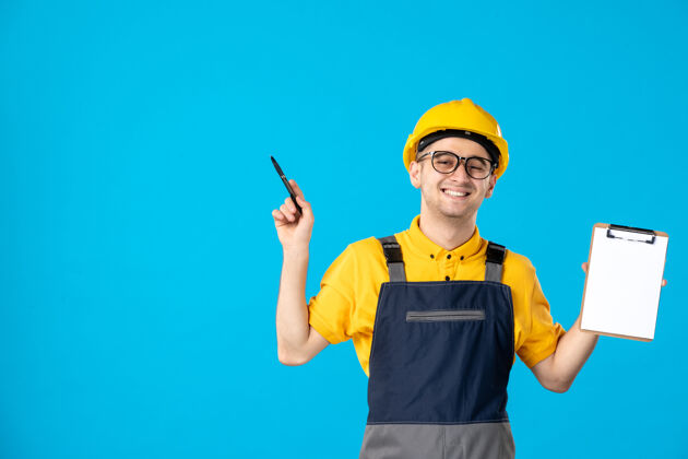 成人身着制服 手拿档案纸条的快乐男建筑工人的正面图在蓝色的墙上文件工作手