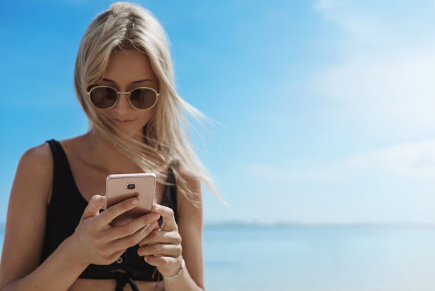 热带迷人的年轻女子在阳光明媚的沙滩上用手机享受暑假比基尼科技海洋