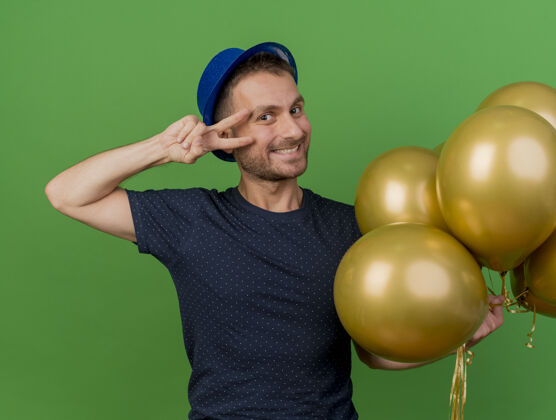 绿色微笑英俊的白人男子戴着蓝色的派对帽 手持氦气球 手势胜利手势 绿色背景上有复制空间标志胜利人