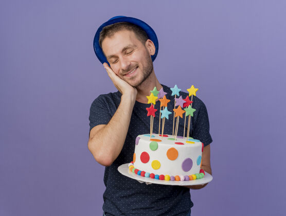 空格戴着蓝色帽子的白人帅哥高兴地把手放在脸上 拿着生日蛋糕 背景是紫色的 上面留有复制空间帽子手紫色