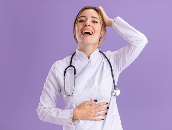 胃笑着的年轻女医生穿着医用长袍 手放在头上 胃被隔离在紫色的墙上穿年轻女