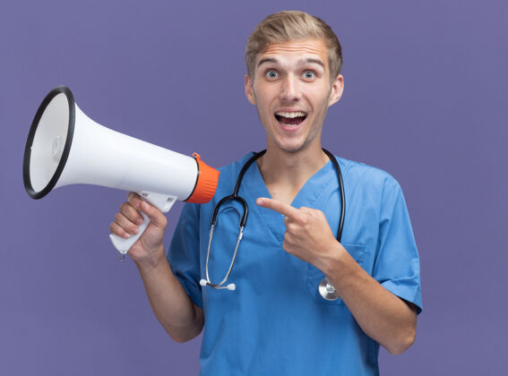 男性微笑的年轻男医生穿着医生制服 手持听诊器 指着隔离在蓝色墙上的扬声器手持制服尖头