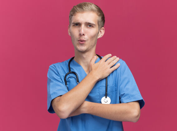 年轻高兴的年轻男医生穿着医生制服听诊器把手放在肩膀上隔离在粉红色的墙上男制服肩膀
