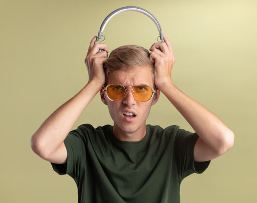 耳机迷茫的年轻帅哥穿着绿色衬衫戴着眼镜头上拿着耳机隔离在橄榄绿的墙上眼镜衬衫头